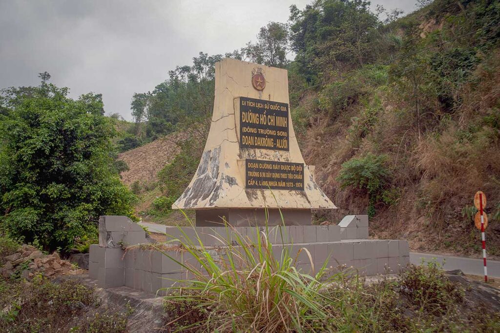Monument marking the Ho Chi Minh Trail in a mountainous area of Vietnam, with the road curving behind it through dense jungle