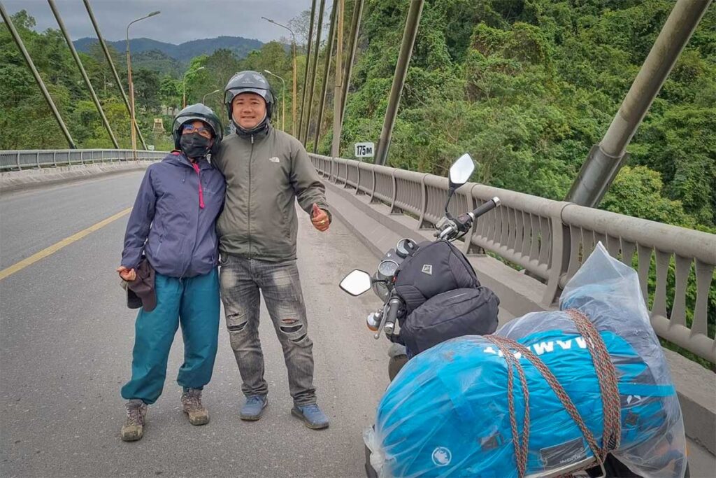 Travelers with an Easy Rider guide on the Ho Chi Minh Road near the DMZ, standing beside a motorbike loaded with luggage on a bridge surrounded by jungle mountains
