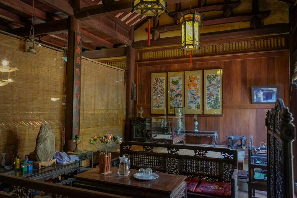 Interior of Duc An Old House in Hoi An featuring antique wooden furniture, hanging lanterns, and hand-painted silk artworks on the wall.