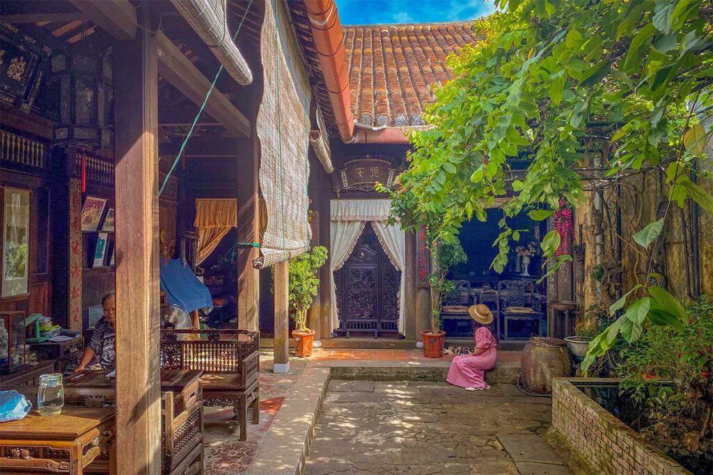 Peaceful courtyard of Duc An Old House in Hoi An with lush greenery, terracotta pots, and a visitor in traditional ao dai sitting under the shade.