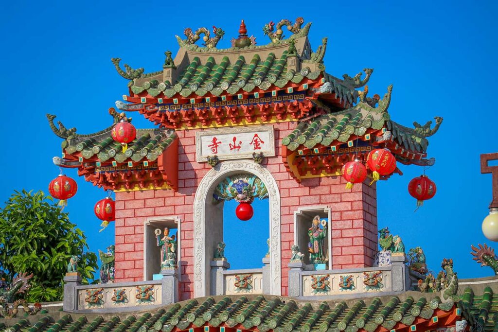 Detailed roof pavilion of Fujian Assembly Hall in Hoi An with green tiles, statues, and red lanterns.