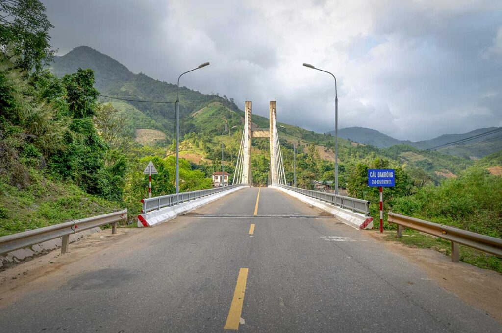 View of Dakrong Bridge on the Ho Chi Minh Road in central Vietnam, with a quiet highway crossing a river and surrounded by green mountains