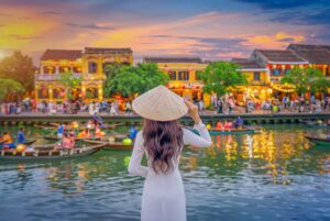 Woman wearing a traditional Vietnamese conical hat overlooking the lantern-lit river and yellow merchant houses of Hoi An Ancient Town at sunset.