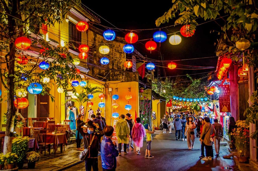 Colorful lanterns illuminating a lively street in Hoi An Ancient Town at night, with people strolling past yellow heritage houses and local shops.