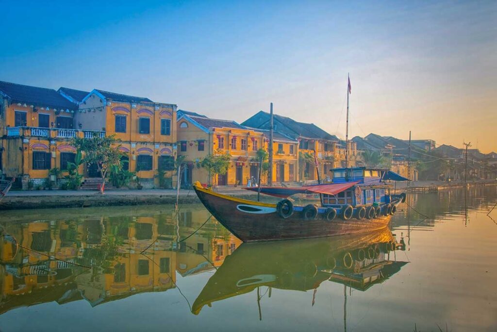 Traditional wooden boat on the Thu Bon River with Hoi An’s yellow heritage houses at sunrise – peaceful morning boat ride in Hoi An.
