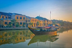 Traditional wooden boat on the Thu Bon River with Hoi An’s yellow heritage houses at sunrise – peaceful morning boat ride in Hoi An.
