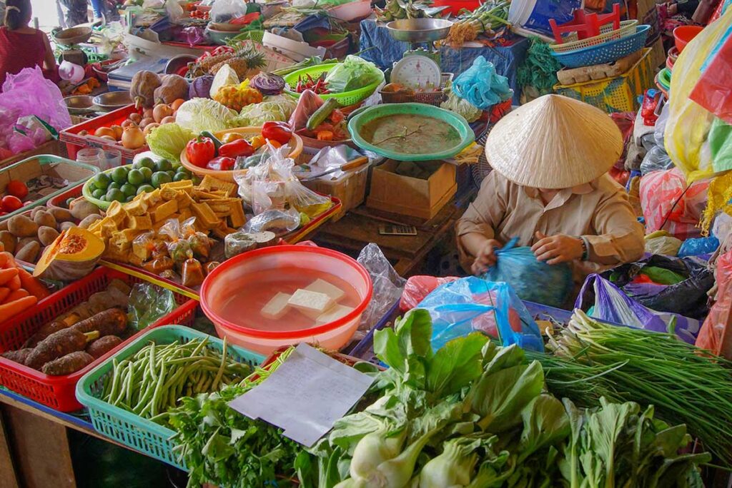 Fresh vegetables at Hoi An Central Market – Colorful display of greens, tofu, limes, and root vegetables sold by a vendor in Hoi An Central Market.
