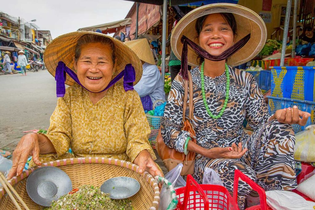 Smiling women at Hoi An Central Market – Two Vietnamese women in conical hats selling fresh herbs with big smiles at Hoi An Central Market, a lively and authentic spot for local produce.