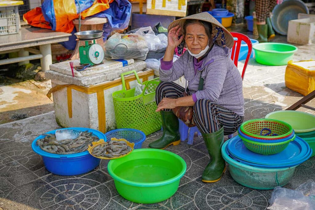 Elderly woman selling shrimp in Hoi An fish market – A vendor in a conical hat sits by tubs of prawns and fresh seafood, reflecting the traditional fishing culture of the town.