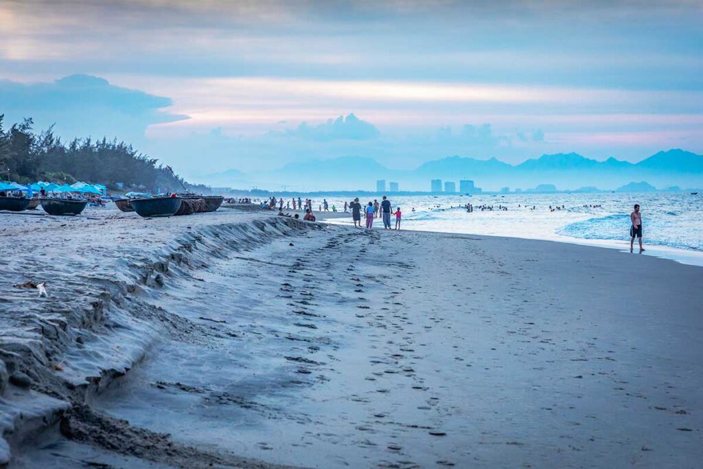 Hoi An Hidden Beach evening walk – Footprints in the sand lead toward the sea as people walk and swim at Hidden Beach near Hoi An, framed by a pastel evening sky and distant mountain backdrop.