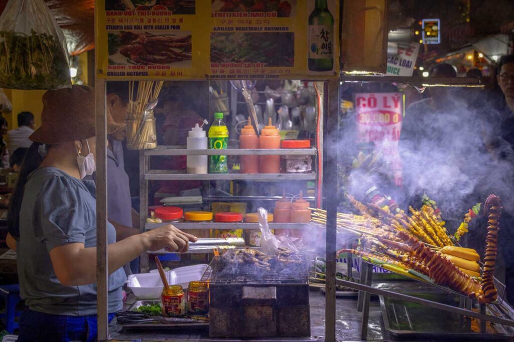Street food vendor grilling octopus and meat skewers at a smoky food stall in Hoi An night market.