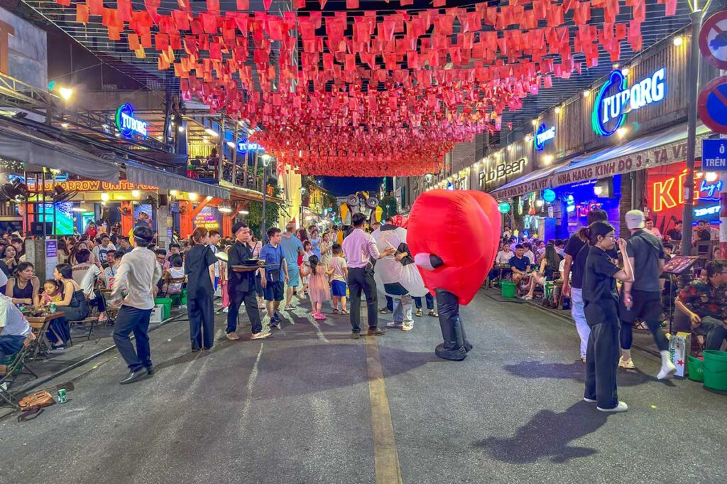Decorated street with flags, a person in a corky costume, locals and foreigners that are all enjoying the atmosphere of Hue Night Walking Street