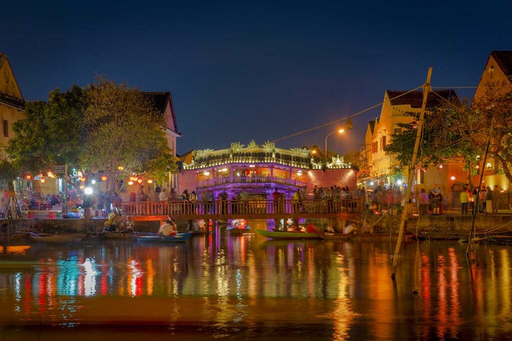 Japanese Bridge Hoi An illuminated at night – A vibrant night view of the Japanese Bridge glowing in colorful lights, reflected in the Thu Bon River as boats pass by during Hoi An’s lantern evening.