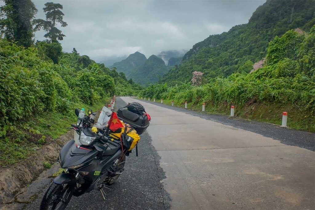 Motorbike parked along the Ho Chi Minh Highway with luggage, overlooking a misty mountain road surrounded by dense jungle