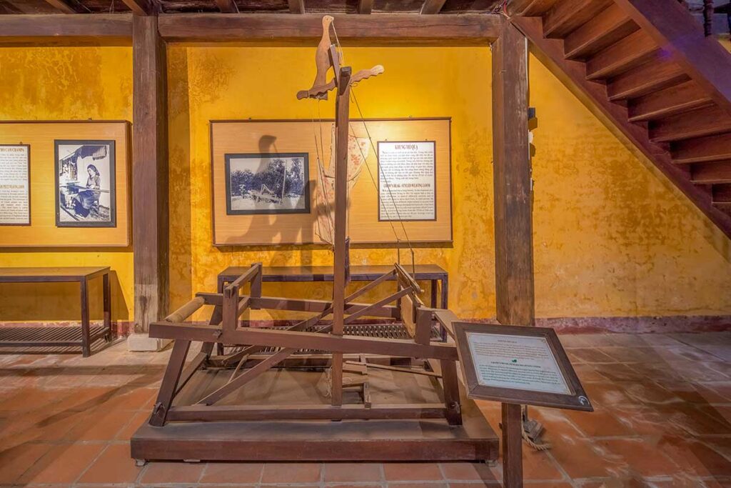 Traditional wooden weaving loom on display at the Museum of Folk Culture in Hoi An, with historic photos and captions explaining local textile craft.”