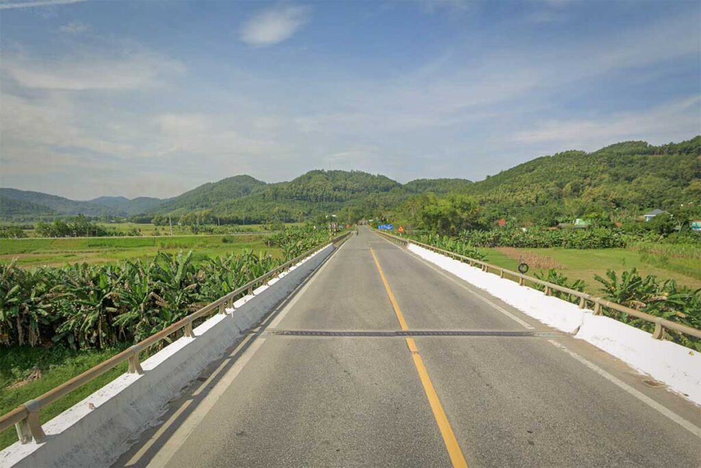 Straight stretch of the Ho Chi Minh Highway through farmland and low hills, with rice fields and banana trees on both sides