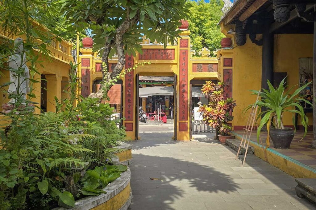 Garden courtyard of Quan Am Pagoda in Hoi An leading to the temple’s main entrance gate – tranquil setting with tropical plants and historic architecture.