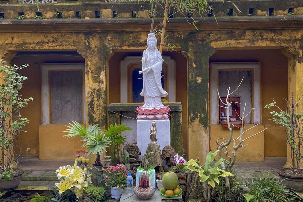 White Quan Âm statue on a lotus pedestal at Quan Âm Pagoda in Hoi An, surrounded by offerings of flowers and fruit in the courtyard.