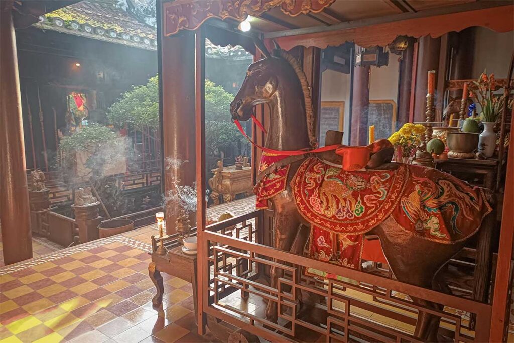 Wooden horse statue inside Quan Cong Temple Hoi An – Sacred wooden horse figure adorned with embroidered red saddle, surrounded by incense smoke in Quan Cong Temple.