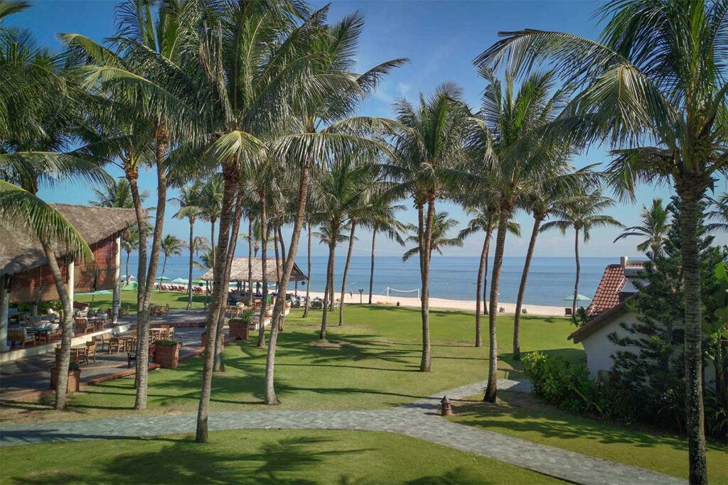 Green lawns and palm-shaded paths leading to the white sands of Cua Dai Beach Hoi An, viewed from a tropical resort.