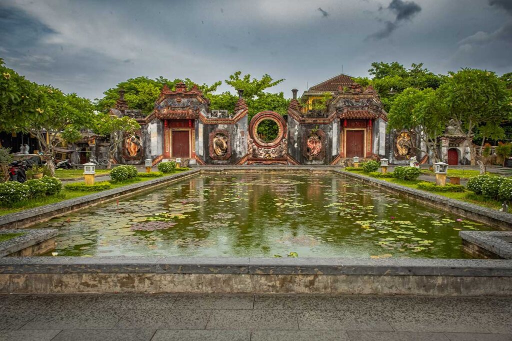 Tâm Ứng Bà Nữ Temple in Hoi An Ancient Town, showing the ancient courtyard, pond, and intricate old stone gates surrounded by greenery.