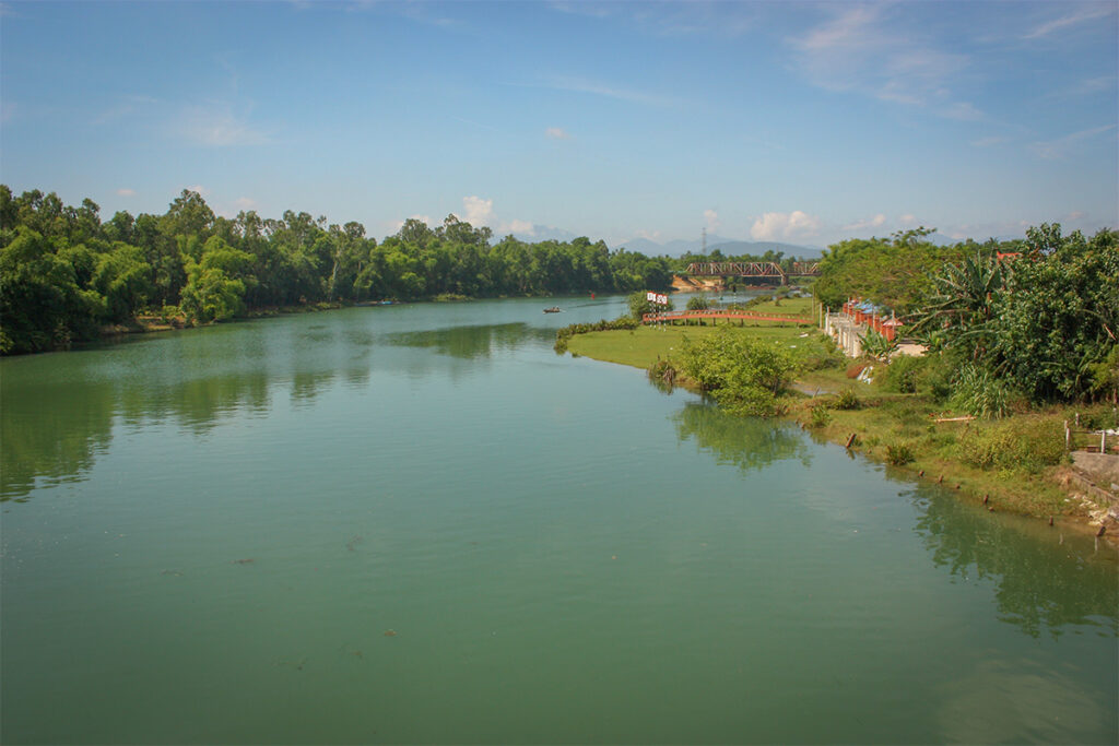 Peaceful view of the Tam Ky River in Quang Nam with green banks, calm water, and a distant bridge under blue sky.