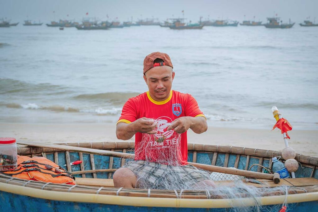 Young fisherman cleaning fishing net inside a basket boat at Tam Tien Fish Market.