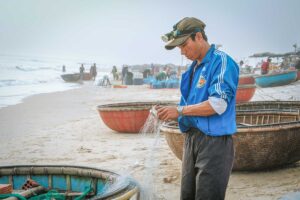 Vietnamese fisherman repairing his fishing net beside a coracle boat at Tam Tien Beach.