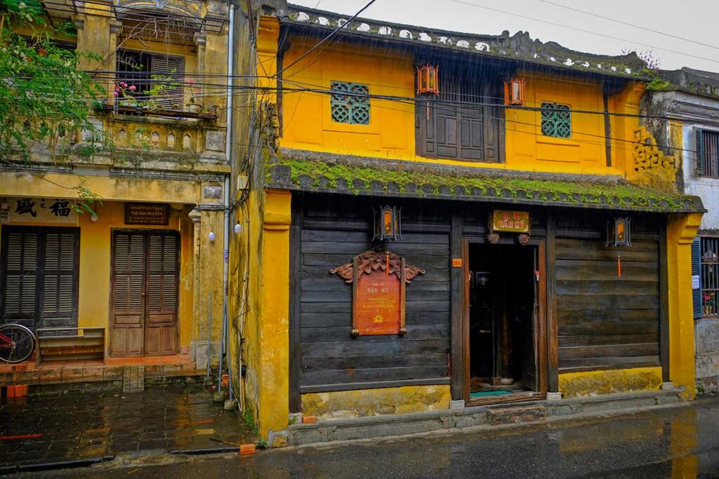 Street view of Tan Ky Old House in Hoi An, yellow facade and black wooden panels with mossy roof tiles on a rainy day.