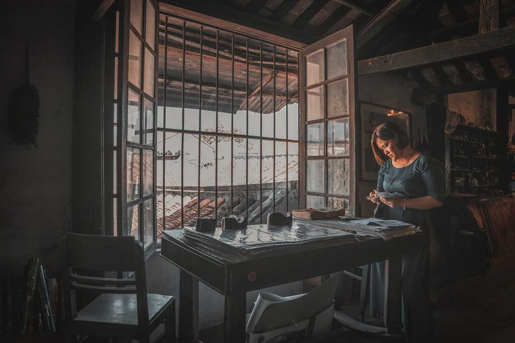 Tourist reading notes by the window of Tan Ky Old House, with wooden beams and historic rooftop views of Hoi An Ancient Town