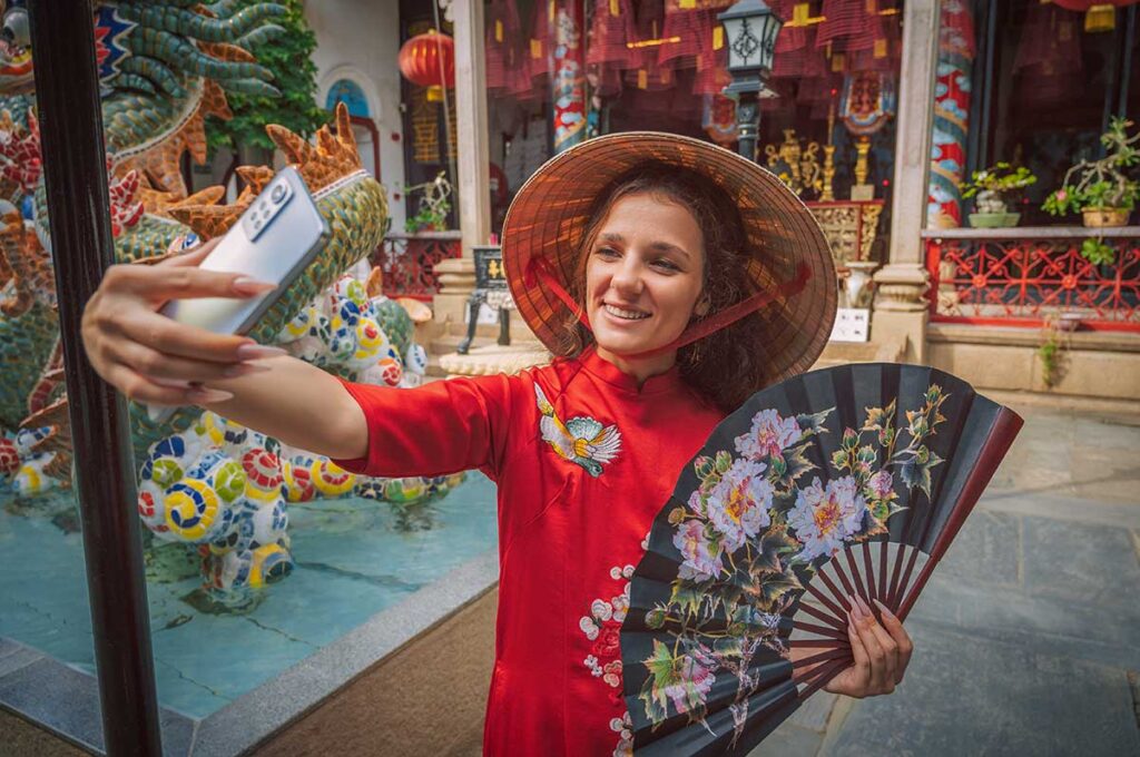 Tourist in traditional ao dai and conical hat taking a selfie at a colorful temple courtyard, showing the cultural and photogenic side of temples in Hoi An.