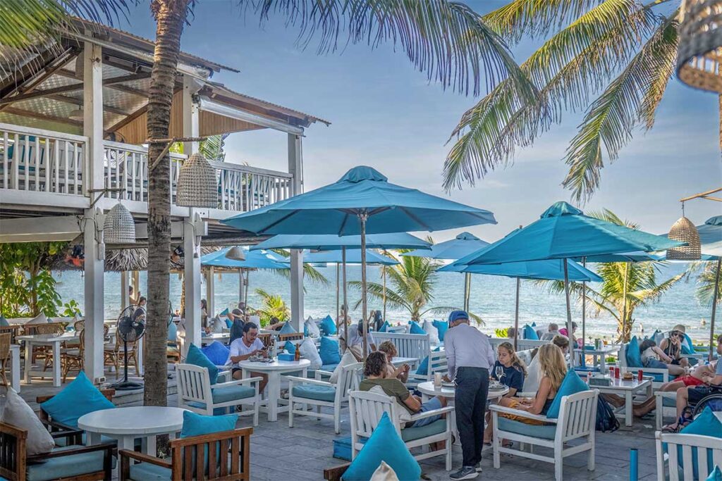 Outdoor dining area of The DeckHouse at An Bang Beach Hoi An, overlooking the sea with turquoise umbrellas and palm trees.