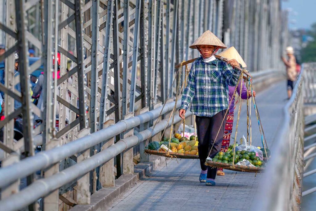 A woman holding a stick with two baskets attached with food inside is walking over the pedestrian side of Trang Tien Bridge