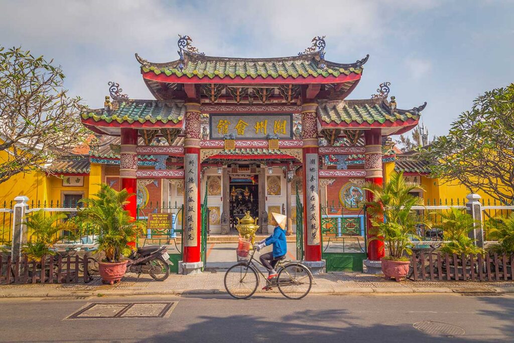 Cyclist passing Trieu Chau Assembly Hall in Hoi An Ancient Town – Daily life scene with a local woman cycling past the colorful Chinese-style temple gate.