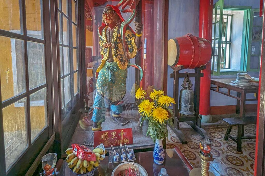 Guardian figure in glass beside offerings of incense, fruit, and flowers inside Trung Hoa Assembly Hall, with temple drum and bell nearby.