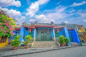 Blue-painted facade of Trung Hoa Assembly Hall in Hoi An, with red lanterns, green iron railings, and Chinese calligraphy.