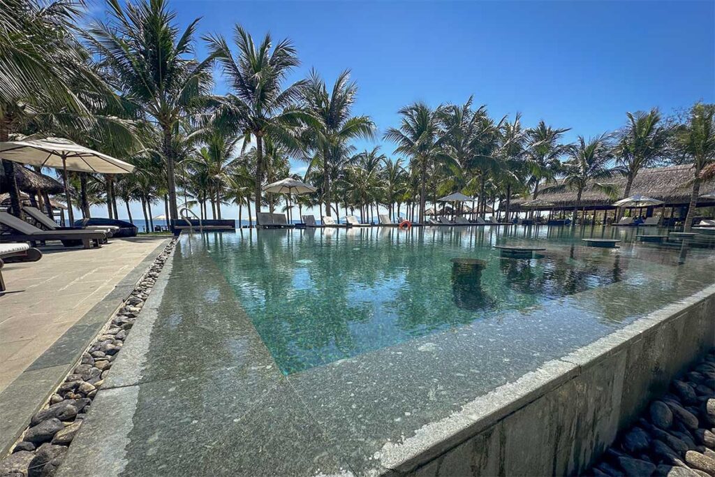 Luxury pool surrounded by palm trees at Wyndham Garden Hoi An near Cua Dai Beach, with sun loungers facing the ocean.