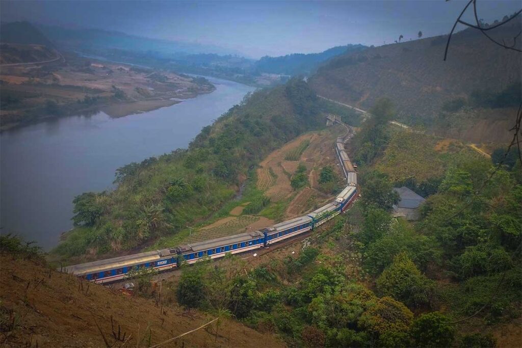 A very long train driving through northern Vietnam landscape along a river making its way from Hanoi to Sapa