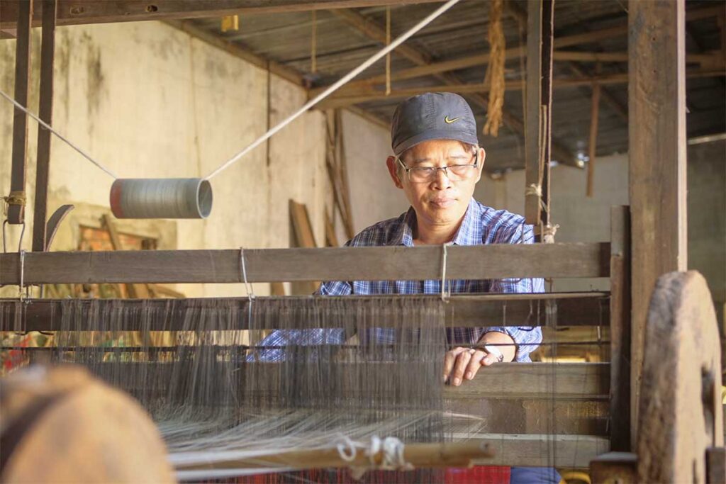 Silk weaver operating a wooden loom in Ma Chau Silk Village, an ancient Hoi An craft village known for handmade silk textiles.