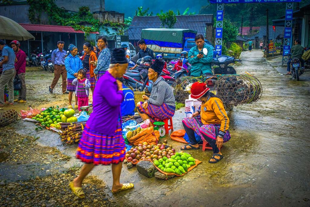 Ethnic woman in traditional clothes is walking past market vendors sitting on the floor selling local produce at the Pa Co Market