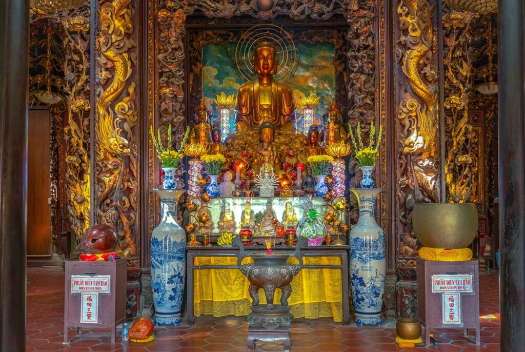 Buddhist altar inside Vinh Trang Pagoda with golden Amitabha statue, lacquered carvings, and offerings of flowers and incense.