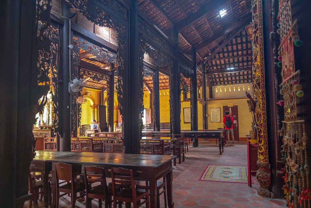 Interior hall of Vinh Trang Pagoda in My Tho, showing dark wooden beams, carved pillars, and traditional Vietnamese temple furniture.