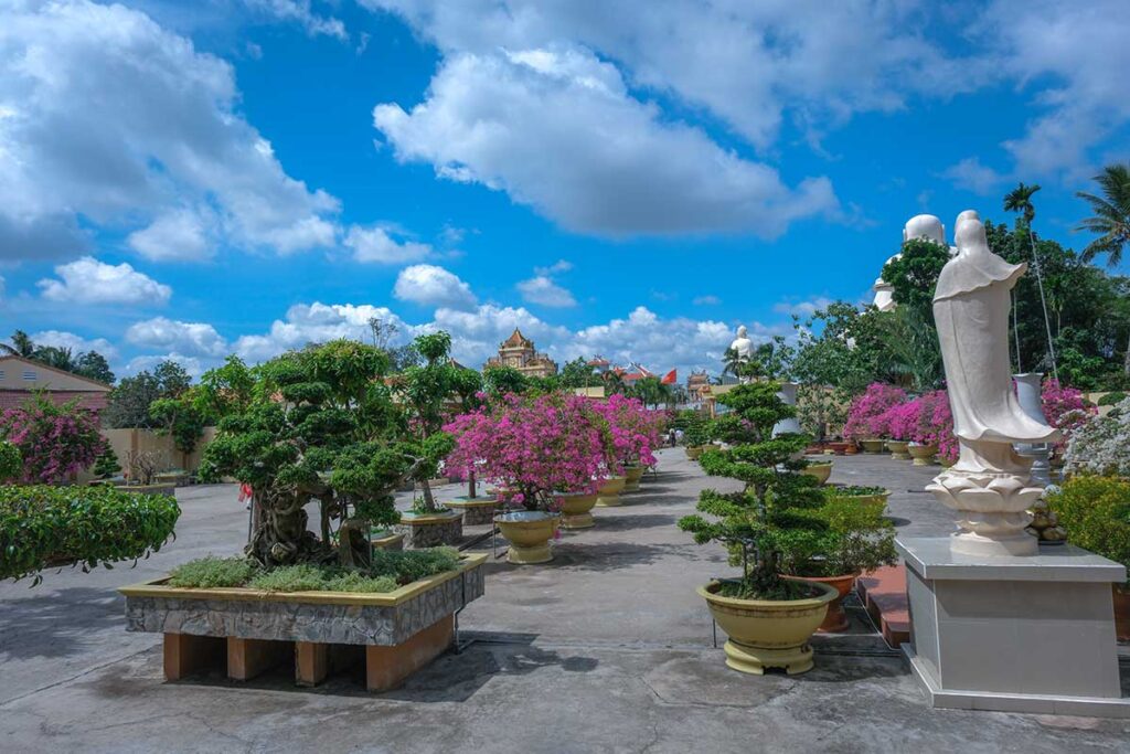 Bonsai garden walkway at Vinh Trang Pagoda in My Tho with flowering trees, statues, and temple buildings in the distance.