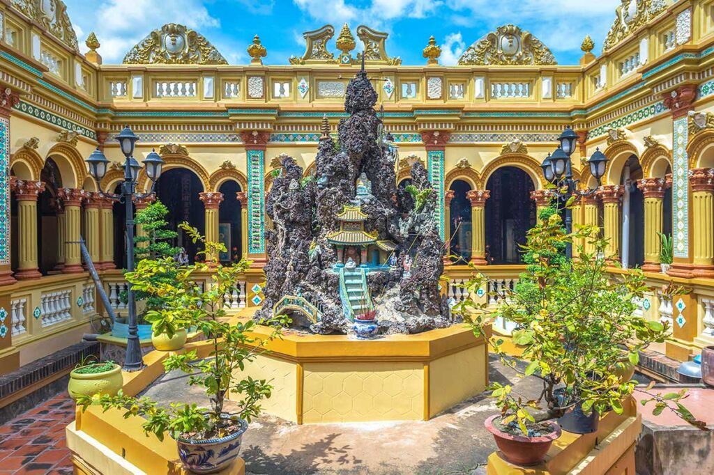 Courtyard of Vinh Trang Pagoda in My Tho with miniature mountain rock garden, bonsai trees, and ornate yellow European-Asian style architecture.