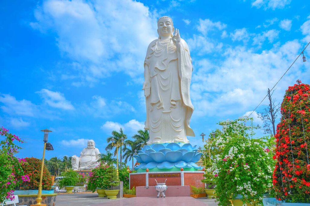 Standing Amitabha Buddha statue at Vinh Trang Pagoda in My Tho, Tien Giang, Vietnam, with Maitreya Buddha in the background and colorful temple gardens.
