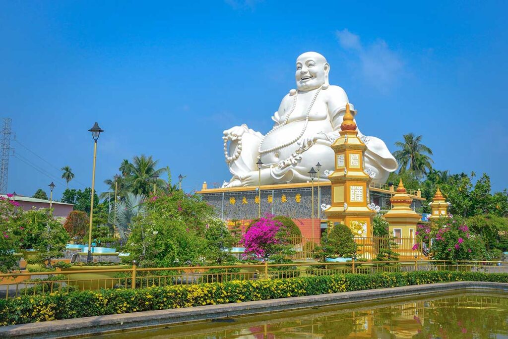 Seated Maitreya Buddha statue at Vinh Trang Pagoda in My Tho, Vietnam, surrounded by gardens, reflecting pond, and bright blue sky.