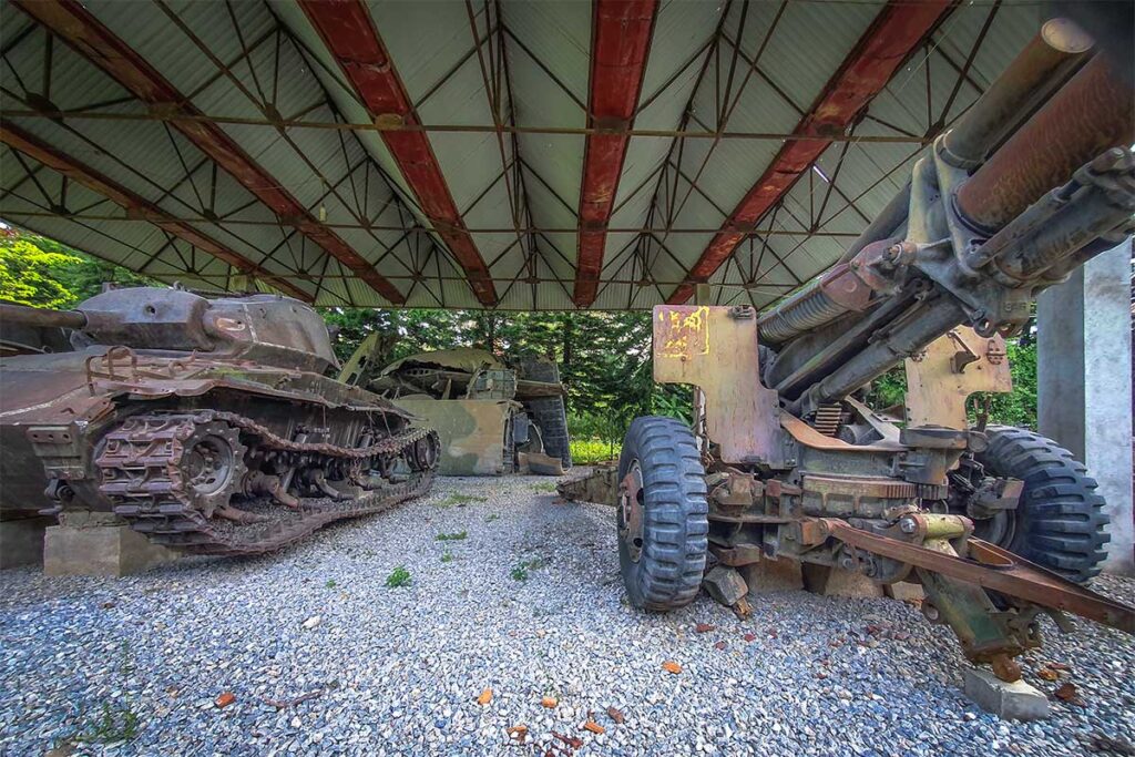 An old rusty canon and tank parked underneath a metal roof as a display at A1 Hill in Dien Bien Phu