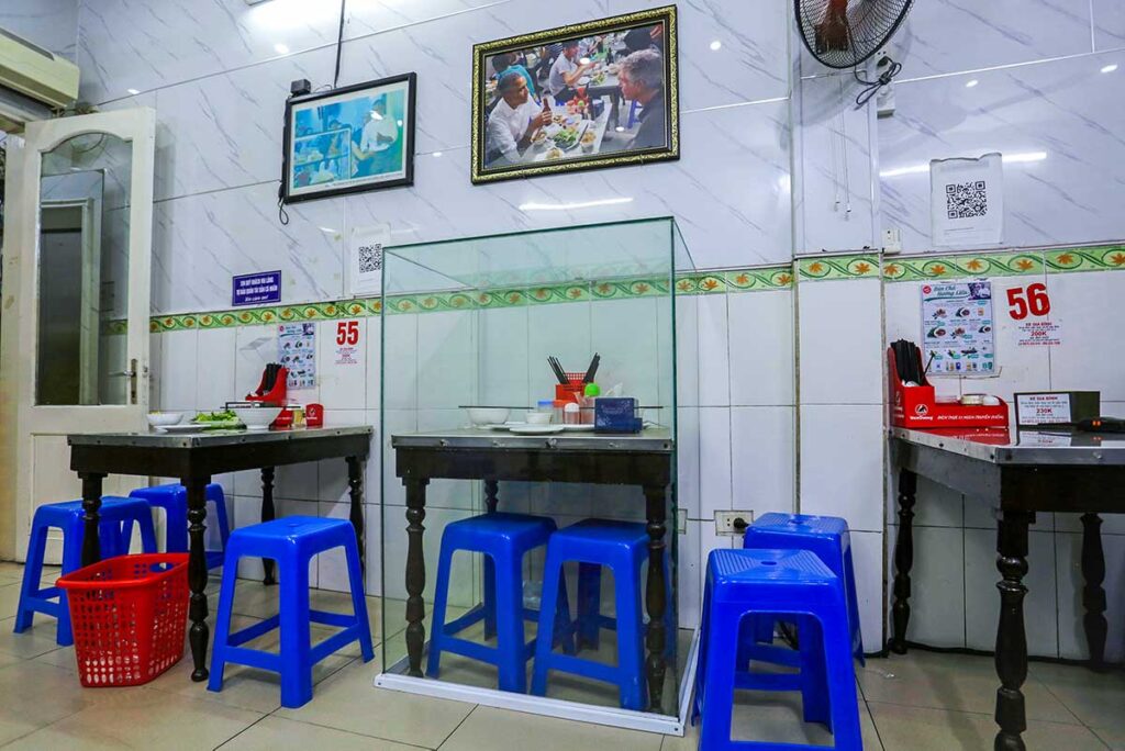 Bún Cha Huong Liên, inside the local Bun Chau Restaurant where a glass structure is set over a table and two plastic chairs where President Obama and Anthony Bourdain ate