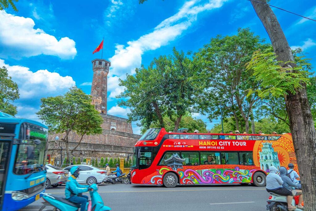 The view from Lenin Park, seen; Hanoi Flag Tower in the background and on the foreground a busy street with motorbikes and the red Hop on Hop Off Hanoi City Tour bus