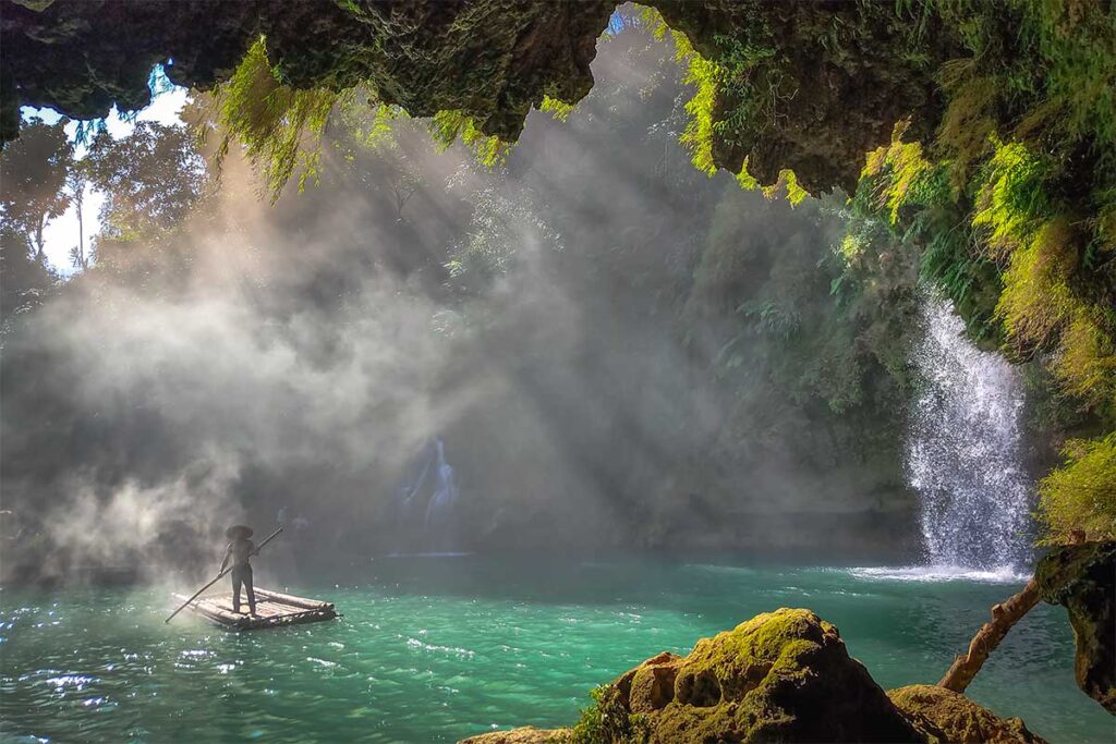 A man standing on a bamboo raft in the mist of Chieng Khoa Waterfall